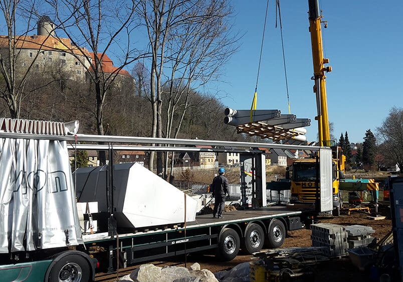 LKW mit Kran entlädt Baustellenmaterial vor Schloss und Häusern, Arbeiter überwacht den Vorgang.