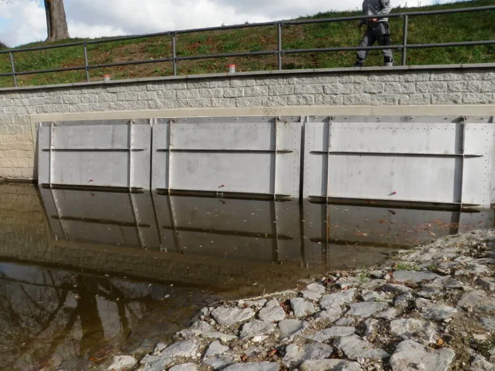 Rückstauklappe Aqua-Flap, Wasserdichtes Hochwasserschutztor aus Metall schützt Uferbereich vor Überschwemmungen, mit einem Gehweg oben.
